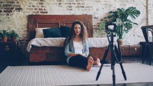 Woman sitting on bedroom floor in front of a camera on a tripod, recording content in a warm brick-walled room