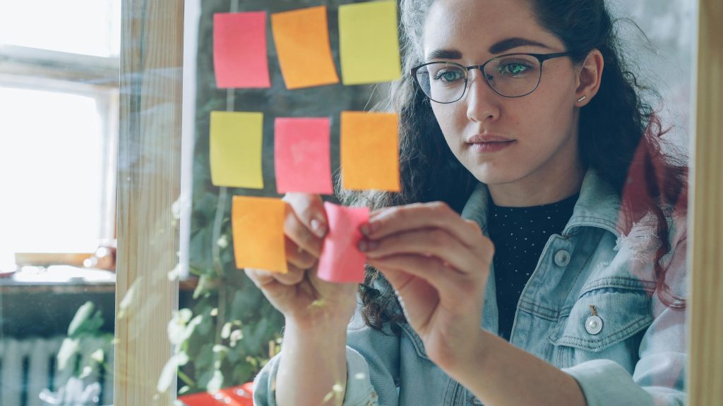 Young woman organising colourful sticky notes on a glass wall to manage share house tasks and responsibilities